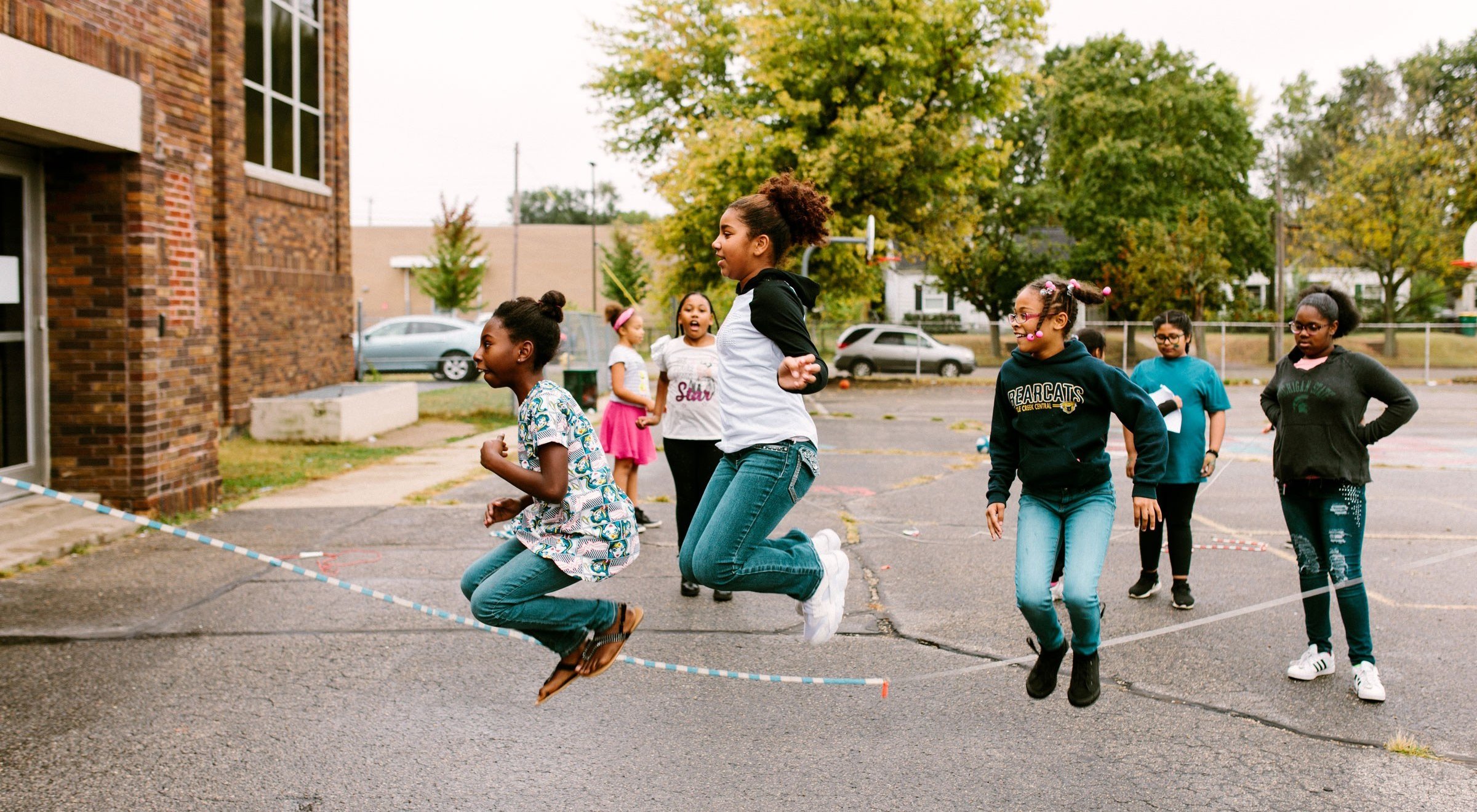 Three students jump rope in the yard while others cheer them on.