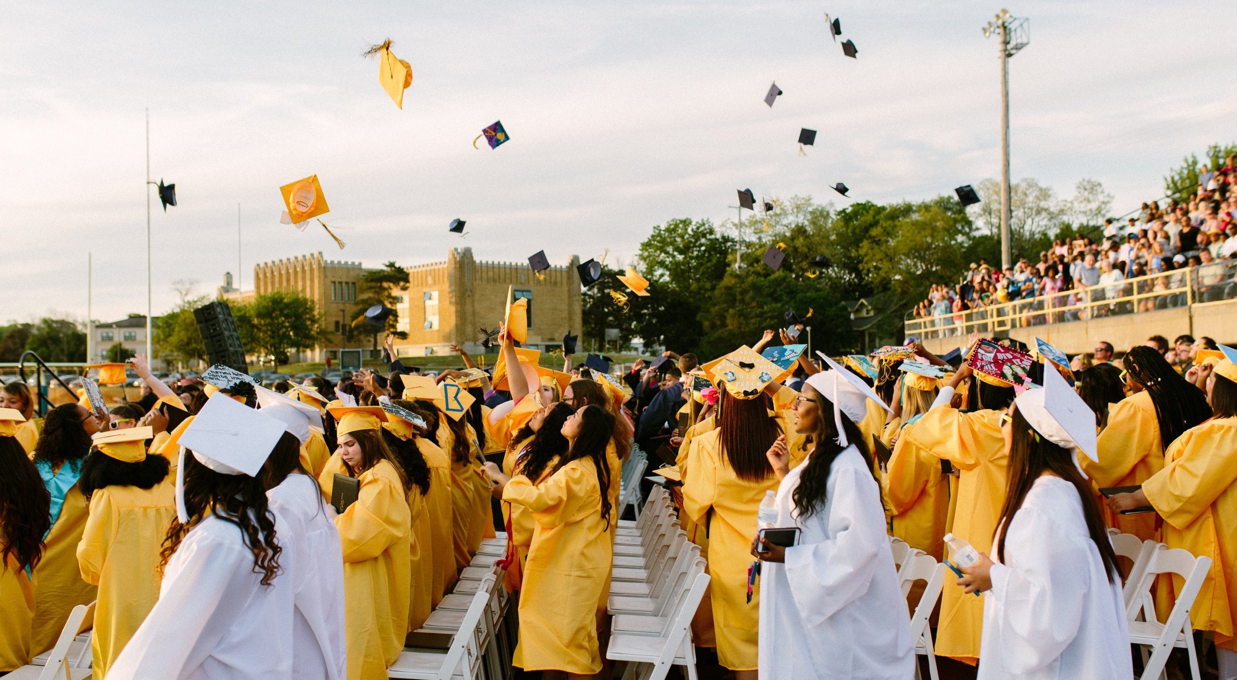 Graduation ceremony with students in yellow and white cap and gown, throwing caps into the air in celebration.